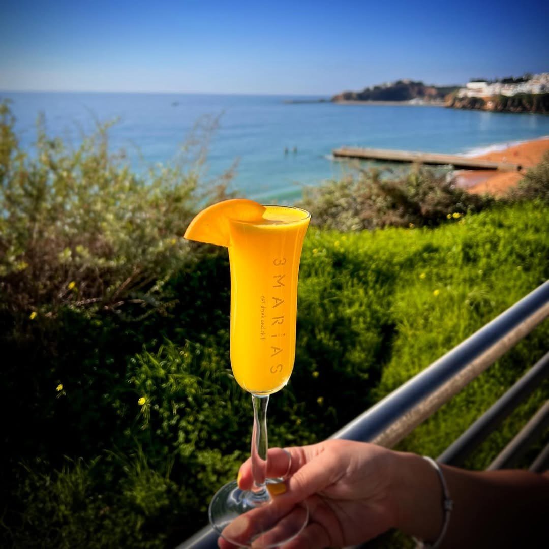 Mão a segurar uma mimosa com rodela de laranja, com a praia e o mar ao fundo